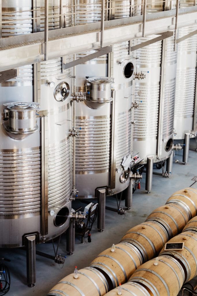 Stainless steel winery tanks beside wooden barrels on a grey production floor